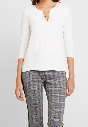 Femme portant un haut blanc à manches trois-quarts avec un col en encoche et un pantalon à carreaux noir et blanc.