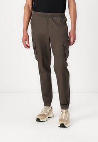 Cargo pants in dark olive green fabric with an elastic waist, side pockets, and tapered legs, paired with beige running shoes.