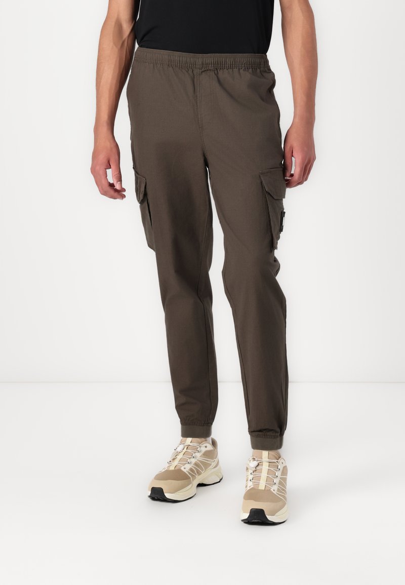 Cargo bukser i mørke oliven grønt stof med elastisk talje, sidelommer og tapered ben, parret med beige løbesko.