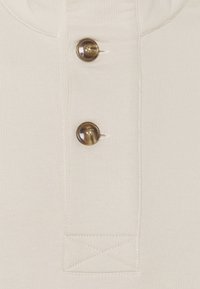 Gros plan sur un tissu beige avec deux boutons en écaille de tortue marron cousus verticalement et des coutures visibles autour d'une pièce rectangulaire.