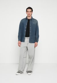 Τζιν πουκάμισο σε σκούρο μπλε με κεντημένο λογότυπο, σε συνδυασμό με ανοιχτό γκρι, φαρδιά τζιν παντελόνια με ραφές και λευκά αθλητικά παπούτσια.