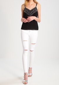 Černý top bez rukávů s korálkovým lemem, doplněný bílými roztrhanými skinny džínami a stříbrnými sandály na podpatku. Jednoduché pozadí.