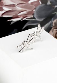 Orecchini a forma di libellula argentata, adornati con pietre trasparenti, con ali dettagliate e un corpo testurizzato, su uno sfondo neutro.