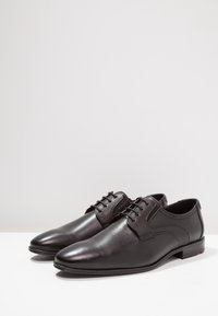 Chaussures habillées en cuir noir avec une finition lisse, un bout rond et des lacets décoratifs, ainsi qu'un panneau texturé sur les côtés.
