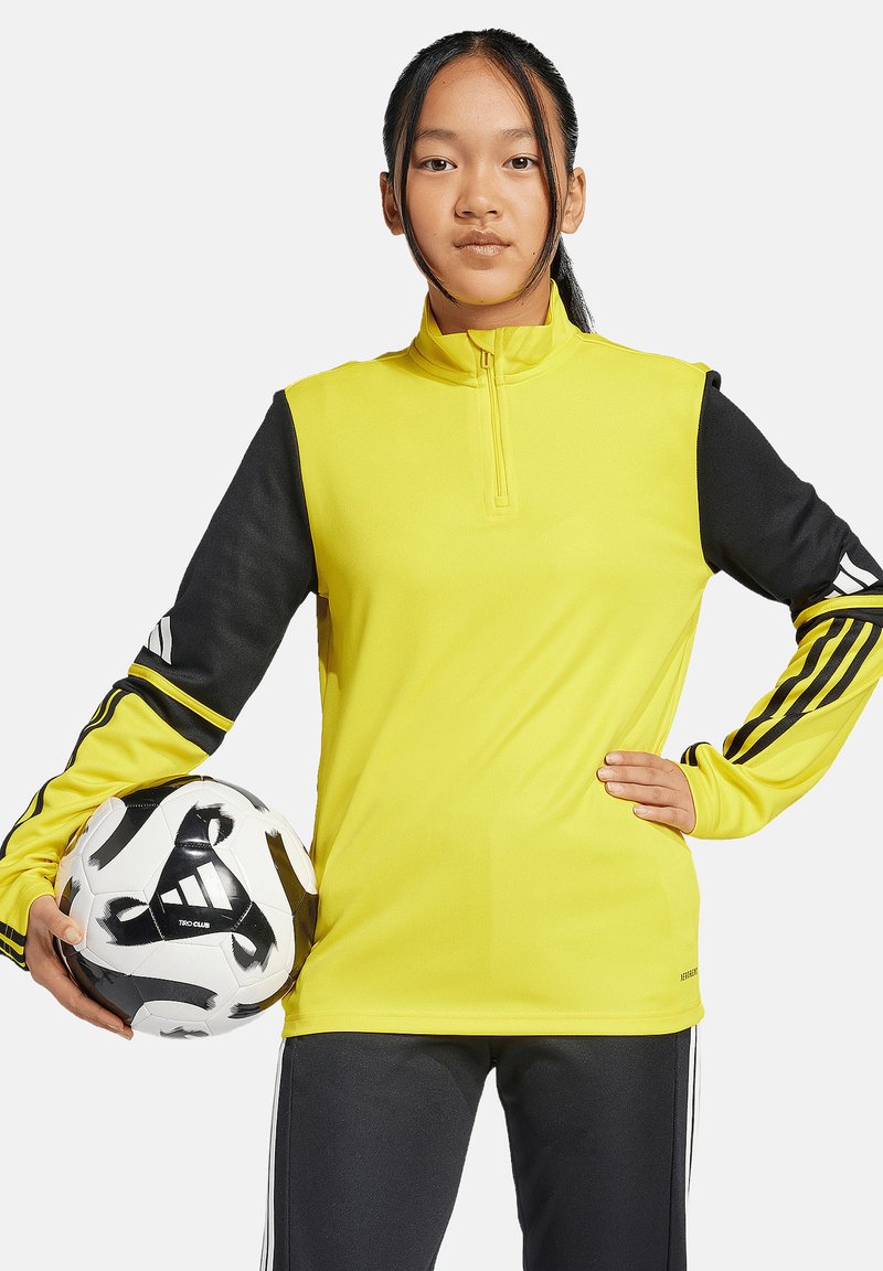 Helles gelbes Sportoberteil mit schwarzen Ärmeln, Reißverschluss am Kragen und drei schwarzen Streifen an den Armen; Model hält einen schwarzweißen Fußball.