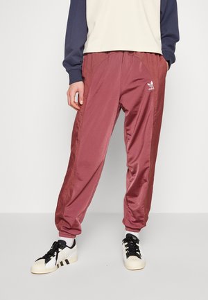 Personne portant un pantalon de survêtement Adidas bordeaux, des baskets blanches avec des lacets noirs, et un haut à manches longues crème et bleu marine, debout devant un fond blanc.