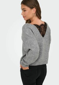 Pull en maille gris à coupe ample, avec un décolleté en V profond au dos, bordé de dentelle noire. Les manches longues et les poignets côtelés complètent le design.