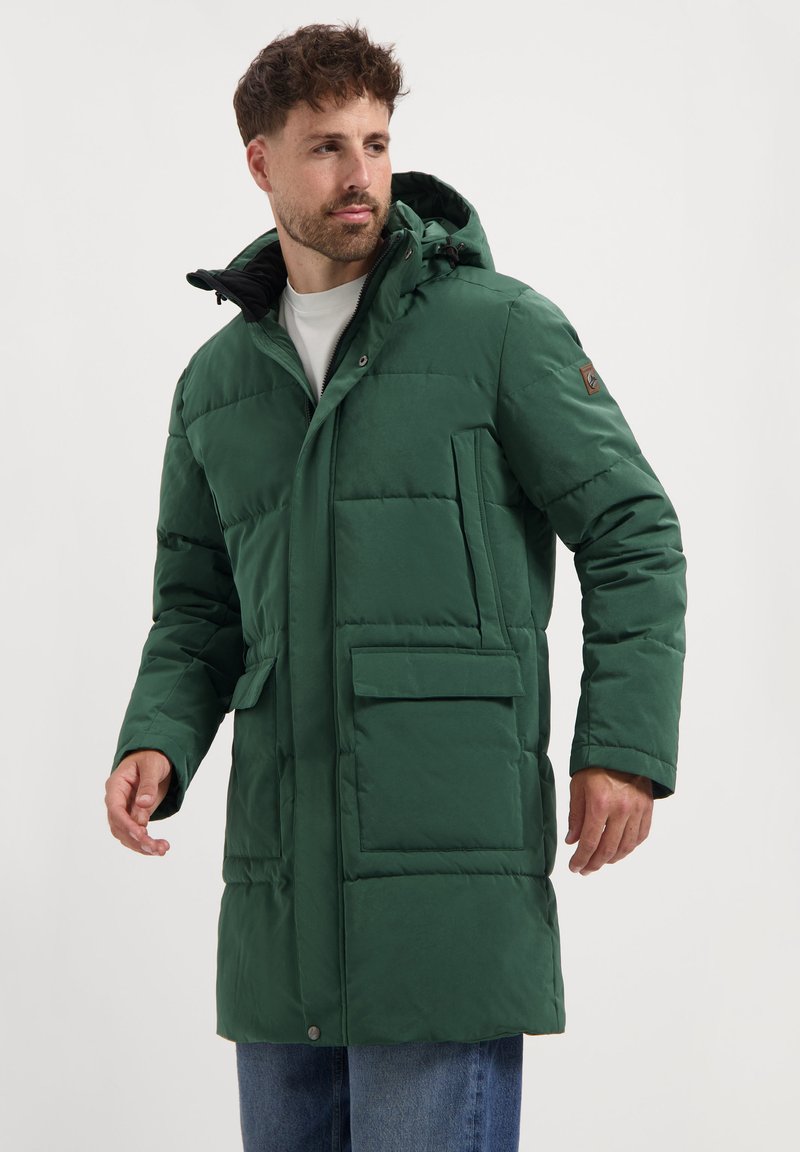 Man in een groene gewatteerde lange parka met capuchon, twee voorzakken met flap en blauwe jeans, kijkt opzij.