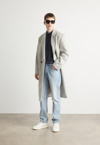 Cappotto di lana grigio chiaro sopra una camicia a stampa scura, abbinato a jeans azzurri chiari e sneakers bianche. Il look è completato da occhiali da sole neri.