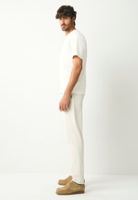 Camicia bianca a maniche corte, pantaloni chiari e scarpe beige in suede. L'outfit presenta un design semplice e pulito con una palette di colori neutri.