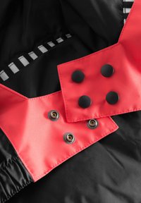 Svart jacka insida med ett klart rött överdrag som har fyra runda svarta tryckknappar och öljetter. Slät textur med kontrasterande element.