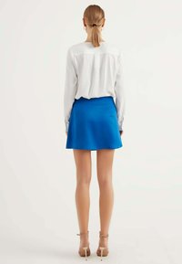 Blå satäng mini kjol i kombination med en vit långärmad skjorta. Slät yta, utsvängd form och ankelremmar på klackskorna synliga.