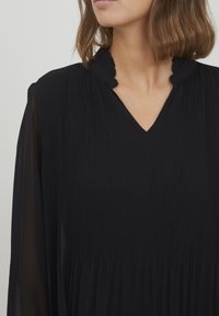 Schwarzes, plissiertes Kleid mit einem gewellten V-Ausschnitt und langen transparenten Ärmeln, das eine fließende Silhouette und eine glatte Textur aufweist.