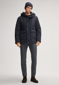 Giacca imbottita blu navy con cappuccio, dotata di due tasche frontali e chiusure a bottone. Abbinata a pantaloni grigi testurizzati e stivaletti marroni. 