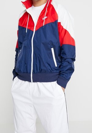 Veste coupe-vent bleu marine, rouge et blanc avec fermeture éclair, poignets élastiques et ourlet. Comprend deux poches avant et un logo sur la poitrine.