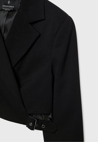 Blazer court noir avec col à revers cranté, manches longues et détail de boucle latérale ajustable, étiquette de marque Stradivarius visible.