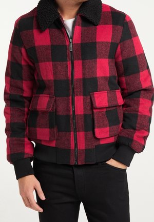 Mand iført en rød og sort ternet lynlåstrøje med en sort lammeskinds-krave, to frontlommer med klap og sorte ribbede manchetter.