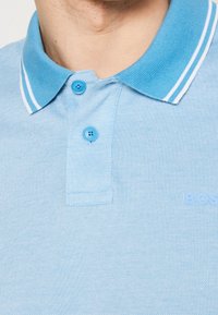 Polo bleu clair en tissu texturé. Il présente une patte de boutonnage, un col contrastant avec des rayures blanches, et un détail de logo en relief.