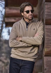 Egy férfi bézs negyed cipzáras pulóverben és napszemüvegben egy fából készült falnak dőlve, összefont karokkal, egy rusztikus szabadtéri környezetben.