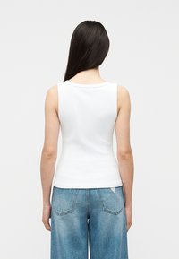 Femme aux longs cheveux foncés portant un haut blanc sans manches et un jean bleu ample, vue de dos sur un fond uni.