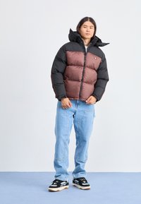 Chaqueta tipo plumón con parte superior negra y sección inferior marrón; presenta una capucha, cierre con cremallera y puños elásticos. Combinada con jeans azul claro y zapatillas negras.