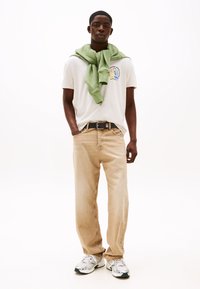 Beige jeans, hvit grafisk t-skjorte, og en grønn genser drapert rundt halsen. Hvite joggesko med grønne aksenter.
