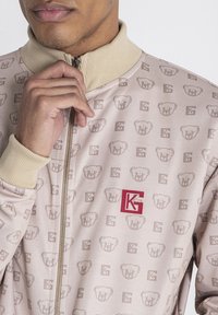 Lichtroze zip-up jack met herhaalde patronen van beren en logo, geribbelde kraag en rode logodetail op de borst.