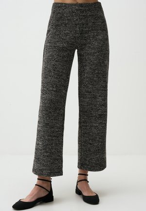 Femme portant un pantalon large en maille côtelée noir et gris avec des chaussures plates noires à bride à la cheville, debout sur un fond clair uni.