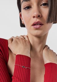 Czerwony, prążkowany top z długim rękawem, z srebrną bransoletką łańcuszkową z ciemnym charms'em i małą czerwoną koraliką; odcień skóry jest ciepły.
