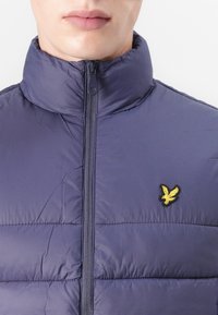 Doudoune marine avec un col haut, dotée d'une fermeture éclair centrale et d'un logo d'oiseau jaune brodé sur le côté gauche de la poitrine.