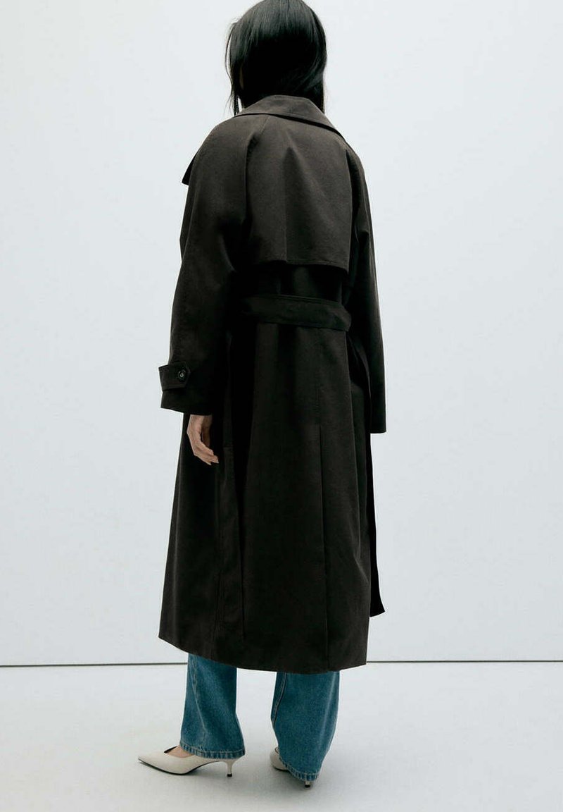 Personne debout de dos, portant un long trench-coat noir, un jean bleu et des chaussures blanches à talons sur un fond uni.