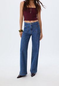 Žena v modrých kalhotách s vysokým pasem a širokými nohavicemi, s předními náprsními kapsami, burgundském zkráceném topu, tmavých špičatých botách a zelenými náramky.