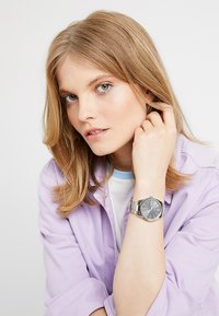 Montre argentée avec un cadran noir rond et des index argentés, portée au poignet. Le modèle a les cheveux châtain clair et est habillé d'une chemise violette.