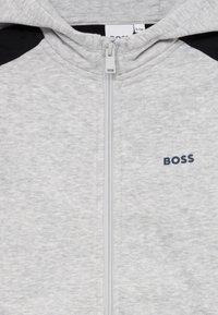Sweatshirt cinzento com fecho éclair, apresentando um tecido texturizado, detalhes pretos nos ombros e um pequeno logótipo azul-marinho no peito.