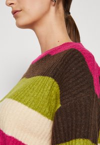 Femme avec une queue de cheval portant un pull en maille côtelée à larges rayures horizontales roses, brunes, vertes et crème.