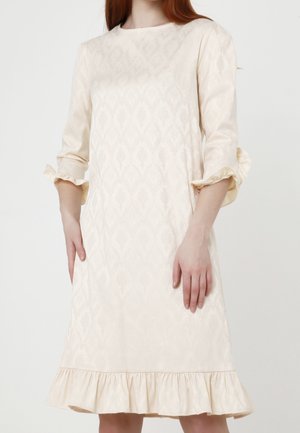 Woman wearing a cream textured knee-length dress with 3/4 sleeves gathered at cuffs and a ruffled hem against a plain background.