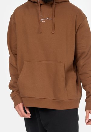 Person trägt einen braunen Hoodie mit Kängurutasche und Kordeln, dazu schwarze Hose, vor schlichtem Hintergrund.