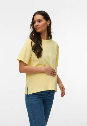 Kvinde med langt mørkt hår iført en løs gul t-shirt og blå jeans, poserer med den ene hånd på maven mod en ensfarvet baggrund.