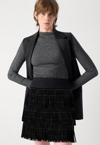 Juoda frakcinė sijonas su metaliniais akcentais, derinamas su sidabriniu raukšlėtu ilgomis rankovėmis marškinėliais, uždengtais juodu be rankovių švarku.