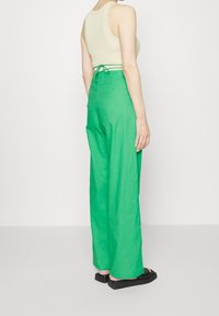 Zielone lniane spodnie o szerokich nogawkach z pasem na sznurku, połączone z beżowym topem na ramiączkach, prezentującym teksturową powierzchnię i luźny krój. Czarne płaskie buty.