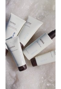 Cinco productos de cuidado de la piel AHAVA están dispuestos sobre una superficie clara y texturizada. Los tubos son blancos con tapas marrones, etiquetados con "AGUA" y los nombres de los productos.