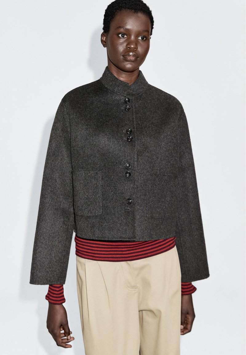 Kobieta nosząca ciemnoszary wełniany żakiet na guziki, pod którym ma na sobie czerwono-czarną w paski bluzkę, w połączeniu z beżowymi spodniami o wysokim stanie.