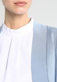 Ljusblå cardigan med ett ribbat mönster, bärs över en vit blus med hög krage. Cardiganen har öppna framkanter och inga knäppningar.