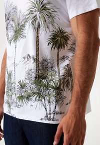 Hvid T-shirt med grøn palmetegning. Korte ærmer, lavet af bomuld. Bæres med mørkeblå jeans, hvilket giver et afslappet look.