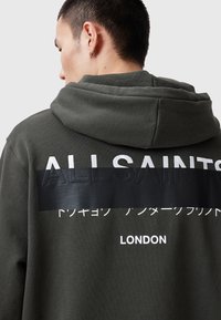 Donkergroene hoodie met een grote, gestructureerde zwarte band met het woord "ALLSAINTS" en Japanse tekst, weergegeven op de achterkant.