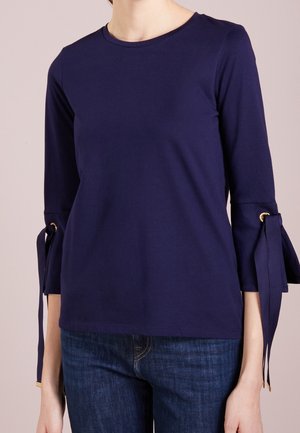 Maglietta a maniche lunghe blu navy con collo tondo, caratterizzata da dettagli a fiocco nei polsi e una texture in cotone liscia. Indossata con jeans di denim.