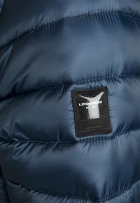 Marinblå pufferjacka med quiltad textur och blankt tyg. Har en svart märkesetikett på vänster sida nära sömmen.