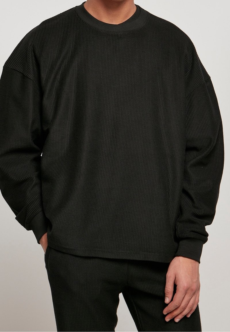 Homme portant un haut à manches longues côtelé noir et un pantalon côtelé noir assorti, une main dans la poche, sur un fond uni.