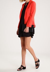 Röd kavaj med långa ärmar över en svart klänning med volangkant, åtföljd av svarta remsandaler och en svart clutch.