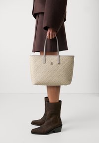 Borsa a tracolla beige con motivo e manici in pelle grigia, con un logo in evidenza. Il modello indossa un abito marrone e stivali marroni abbinati.